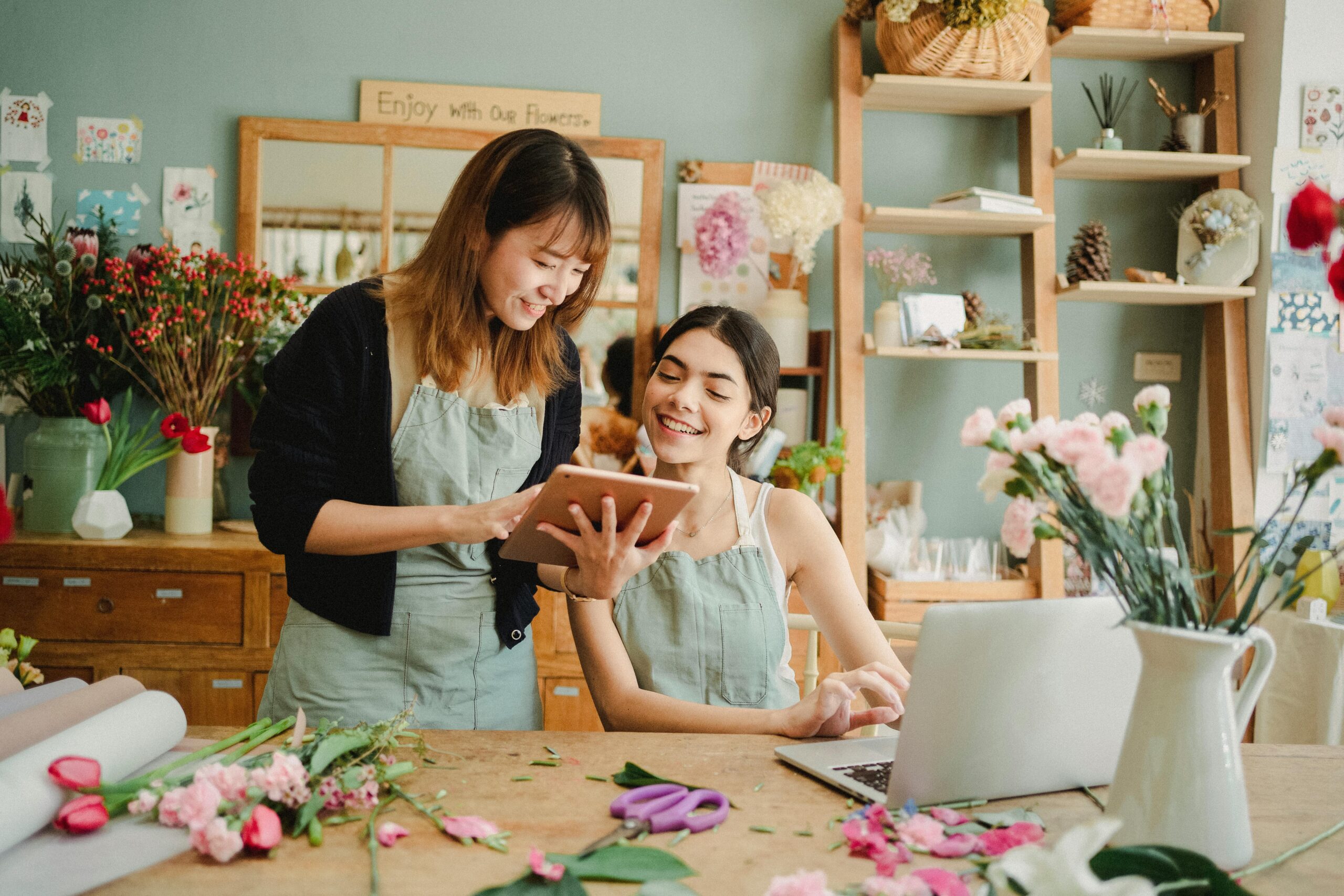 In a flower shop with pale teal walls, an asian woman with shoulder-length hair shows a white woman with a ponytail information on a tablet.